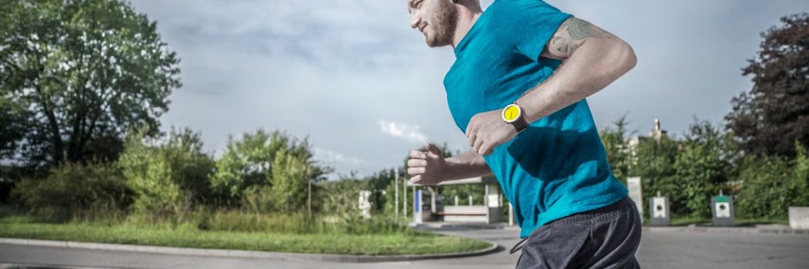 Caucasian man jogging on a city street crosswalk wearing a blue t-shirt and yellow watch.