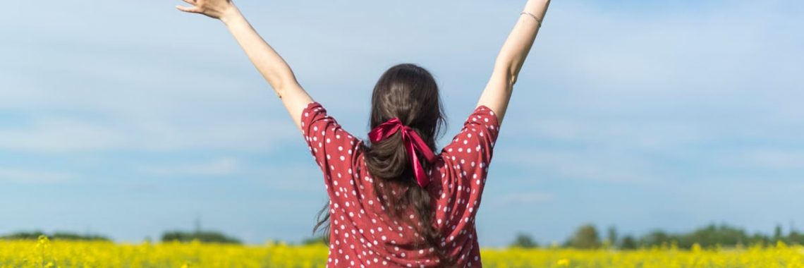 A woman in a polka dot shirt raising her arms in a bright yellow flower field, expressing freedom and joy.